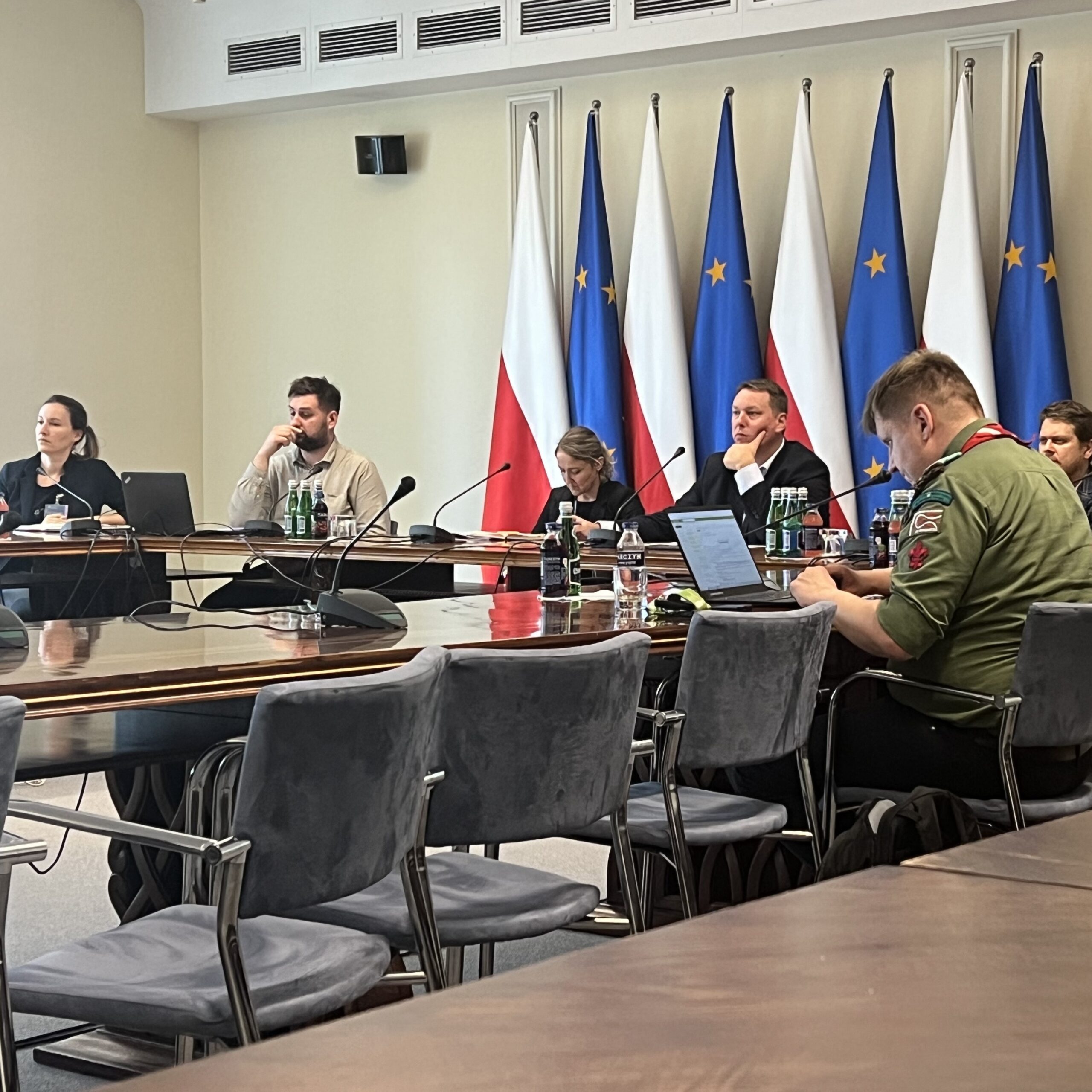 Ludzie siedzący przy stole konferencyjnym na tle flag Polski i UE.