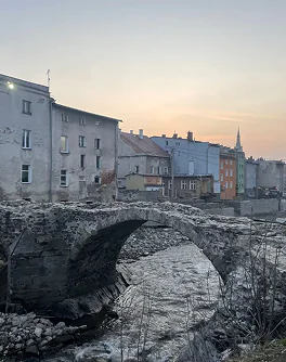 Kamienny most nad strumieniem z budynkami w tle o zachodzie słońca.